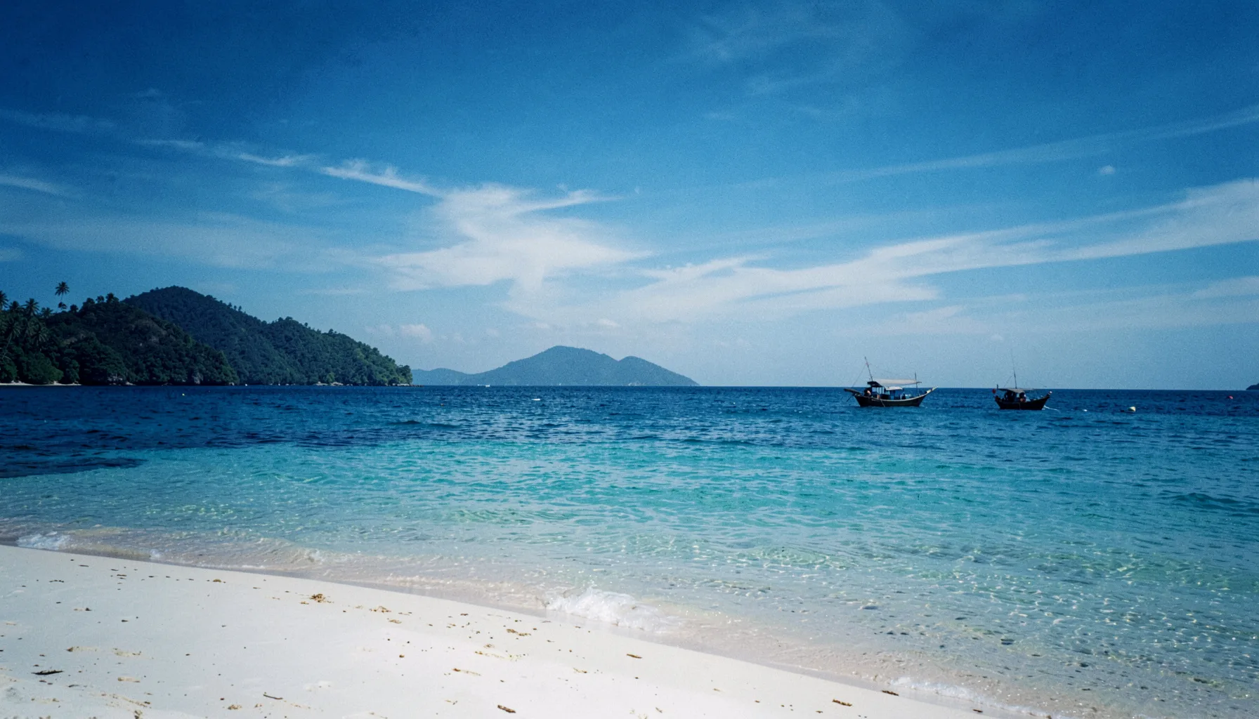 Pemandangan pantai Lombok dengan pasir putih, air laut biru jernih, dan perahu tradisional di kejauhan, langit cerah dengan awan tipis.