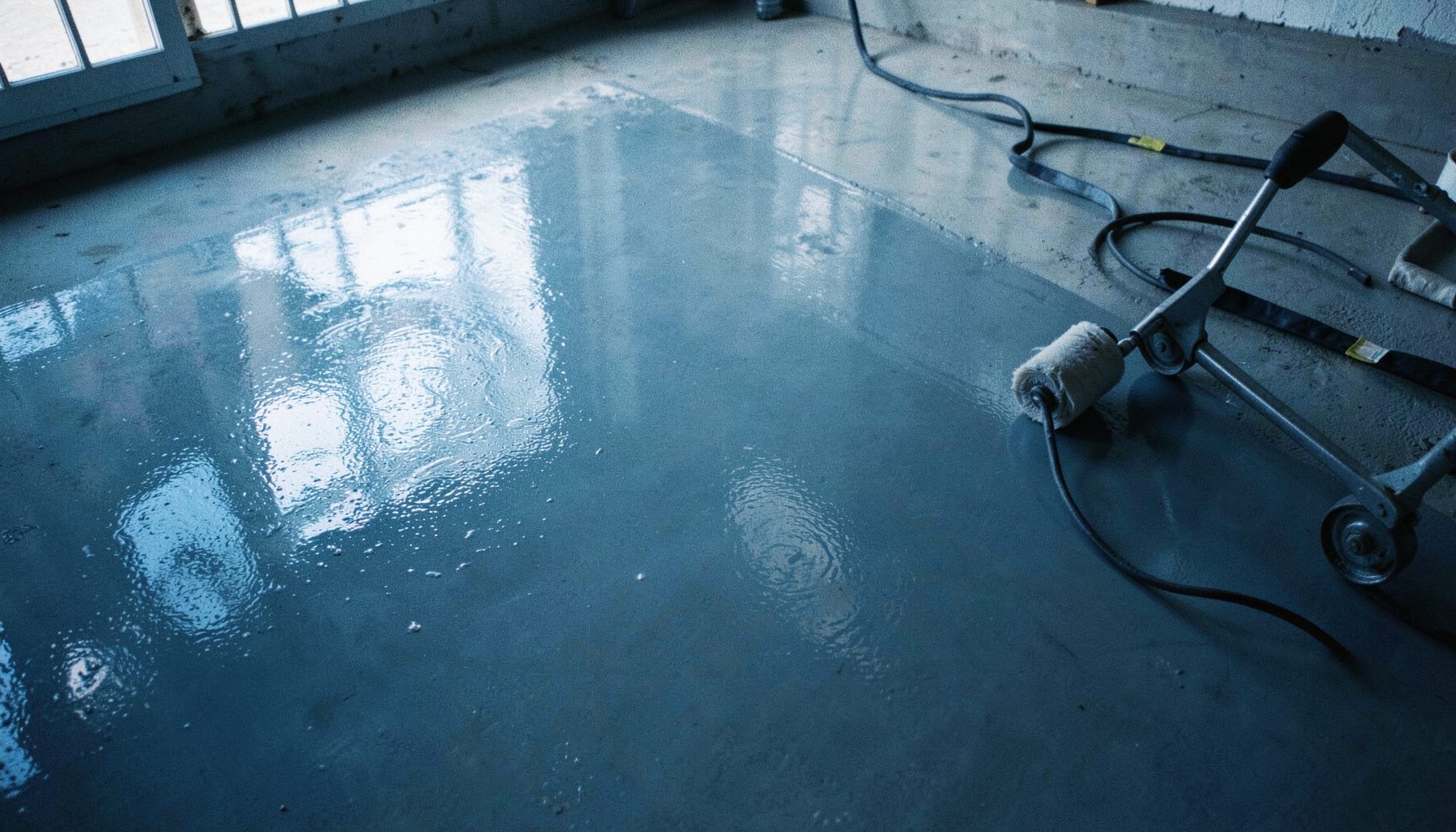 Lantai beton abu-abu yang sedang dilapisi epoxy mengkilap, dengan rol dan alat aplikasi di sampingnya